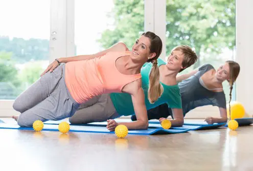 Drei Frauen führen gemeinsam eine seitliche Plank-Übung in einem hellen Kursraum durch, um ihre Rumpfmuskulatur und Stabilität zu stärken.