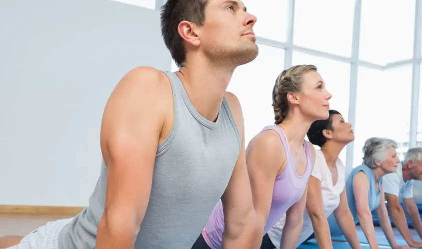 Eine Gruppe von Menschen unterschiedlichen Alters führt gemeinsam Yoga-Übungen in einem hellen Kursraum durch, um Beweglichkeit und Wohlbefinden zu fördern.