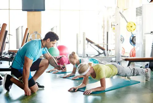 Eine Gruppe von Männern und Frauen die durch Anleitung eines Trainers gemeinsam eine Plank-Übung in einem Fitnesskurs ausführt, um ihre Kraft und Stabilität des Rückens zu trainieren.