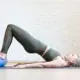 Eine Frau führt auf einer Yogamatte eine Beckenbrücke mit Unterstützung einer Schaumstoffrolle durch, um ihre Rumpfmuskulatur und Stabilität zu trainieren.