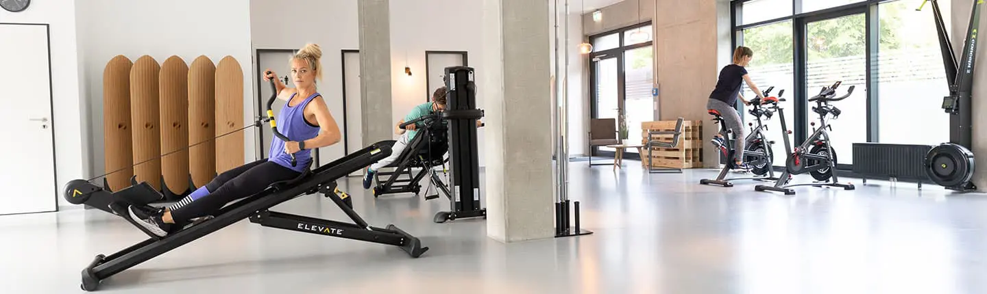 Personen trainieren in einem modernen Fitnessraum mit verschiedenen Geräten, darunter ein Rudergerät, Kraftgerät und Fahrradergometer, zur Förderung von Kraft und Ausdauer.